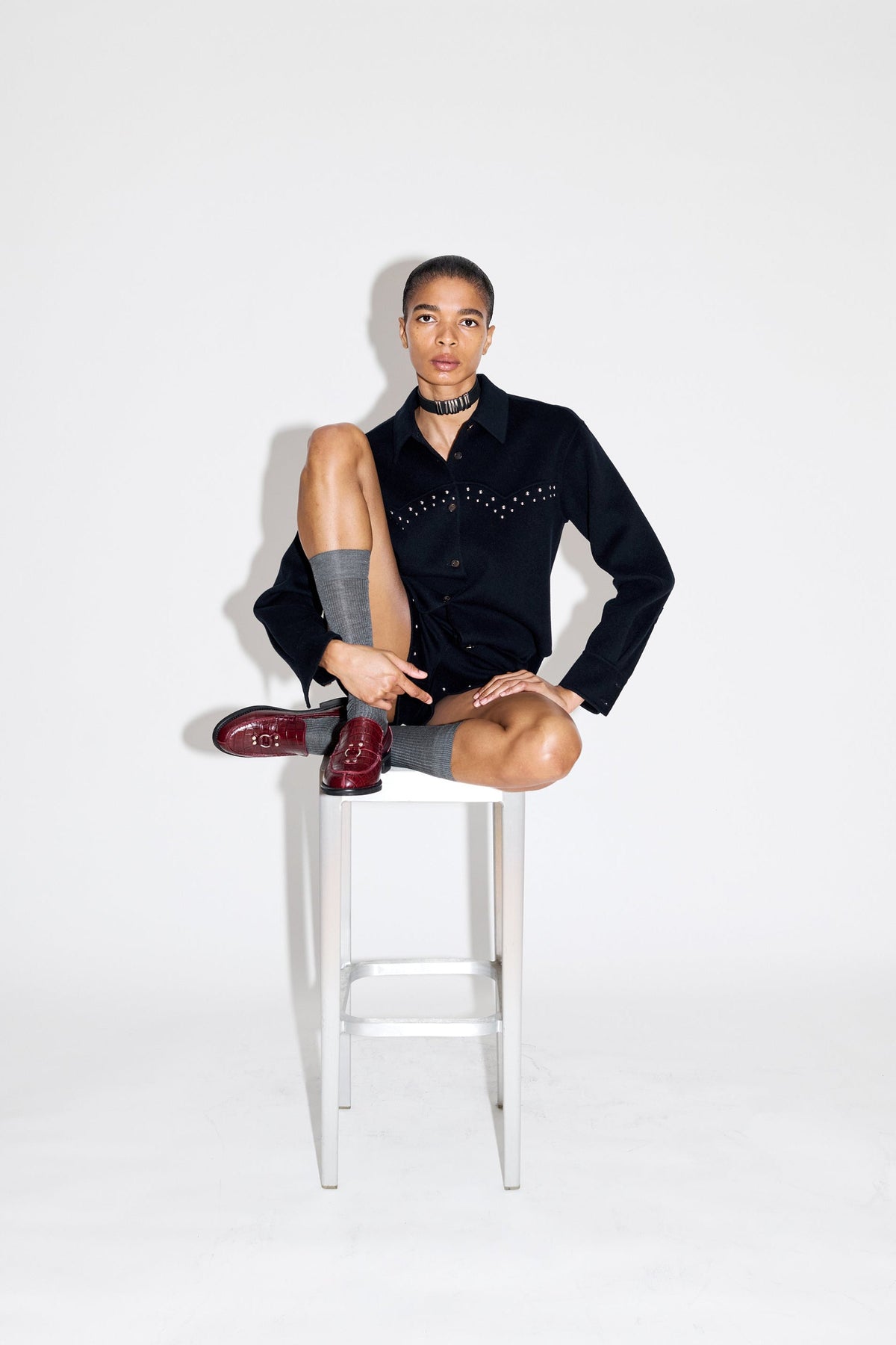 A model sits on a white metal stool against a white studio background, wearing a black long-sleeve button-up shirt with silver stud embellishments on the chest, gray socks, and burgundy dress shoes.