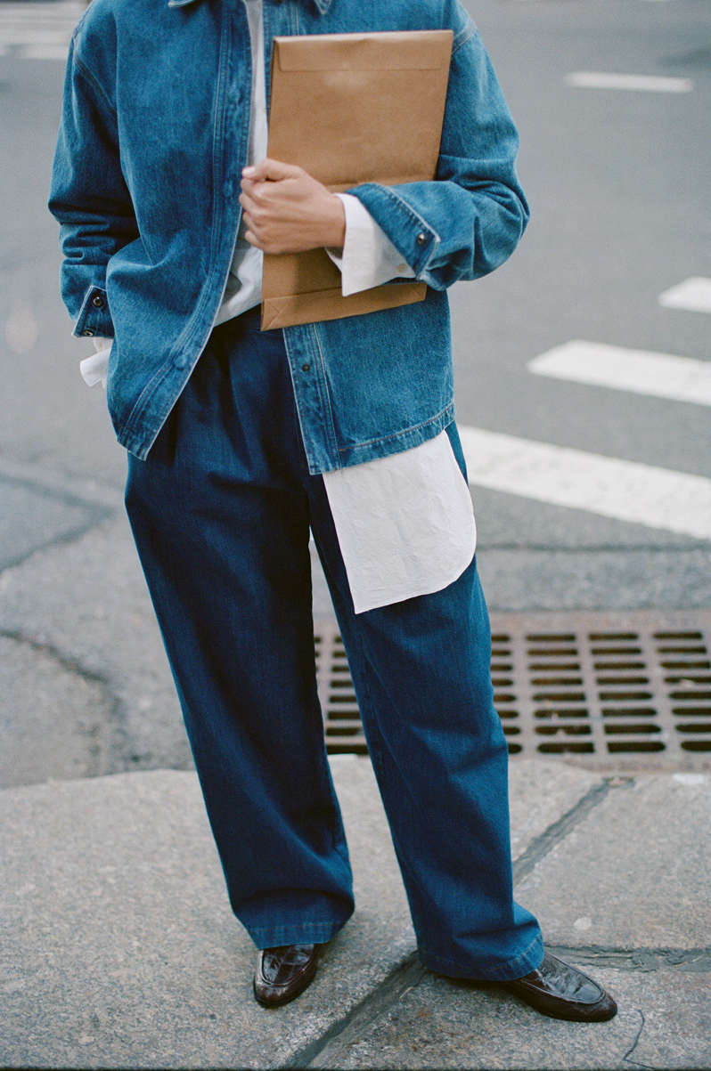 Person wearing a blue denim jacket and pants holding a brown book on a street.