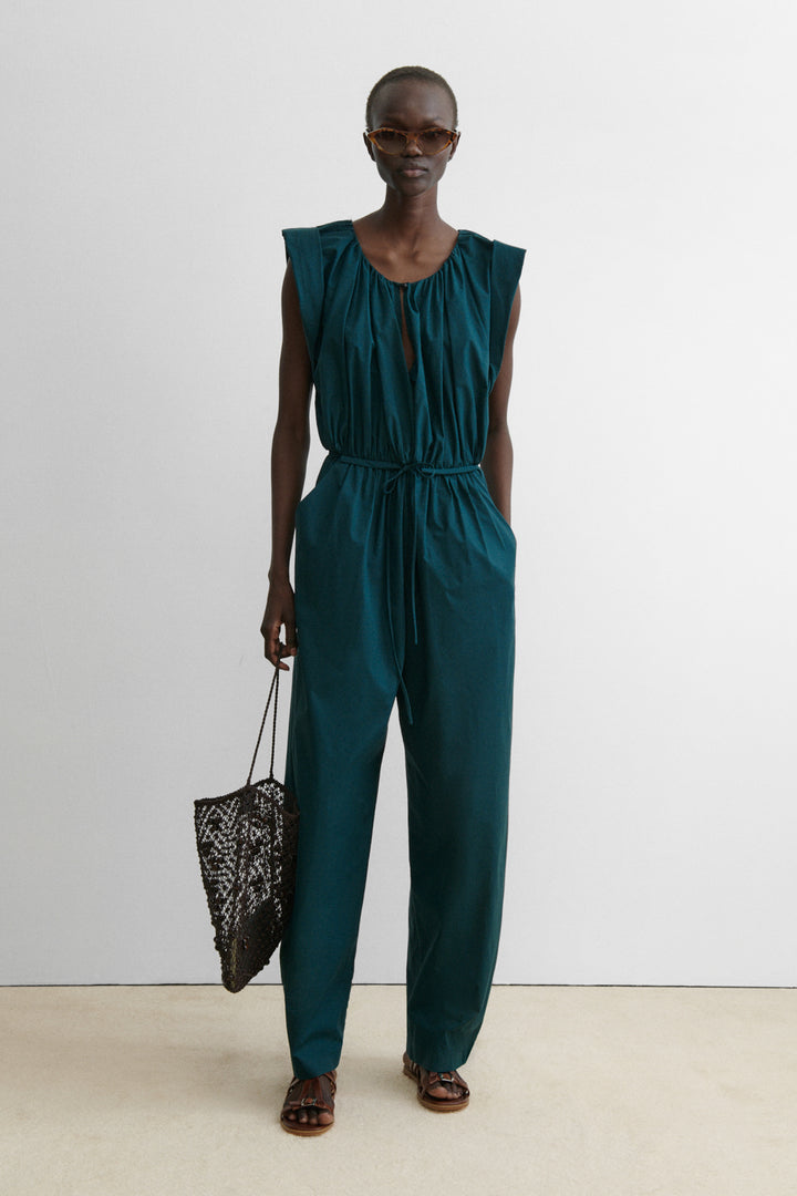 Photo of a model in sunglasses holding a mesh bag and wearing a short-sleeved, dark teal gathered jumpsuit