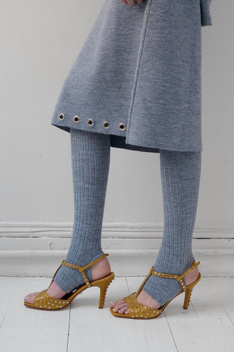 A person wearing a light blue-gray knit dress, matching ribbed tights, and gold heeled sandals with metallic studs, standing against a plain off-white wall and white floor.