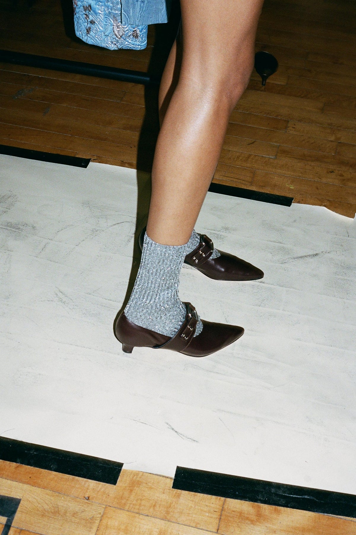 A person's legs wearing brown pointed-toe Mary Jane shoes with a silver buckle and gray ribbed socks, standing on a white sheet over a wooden floor.