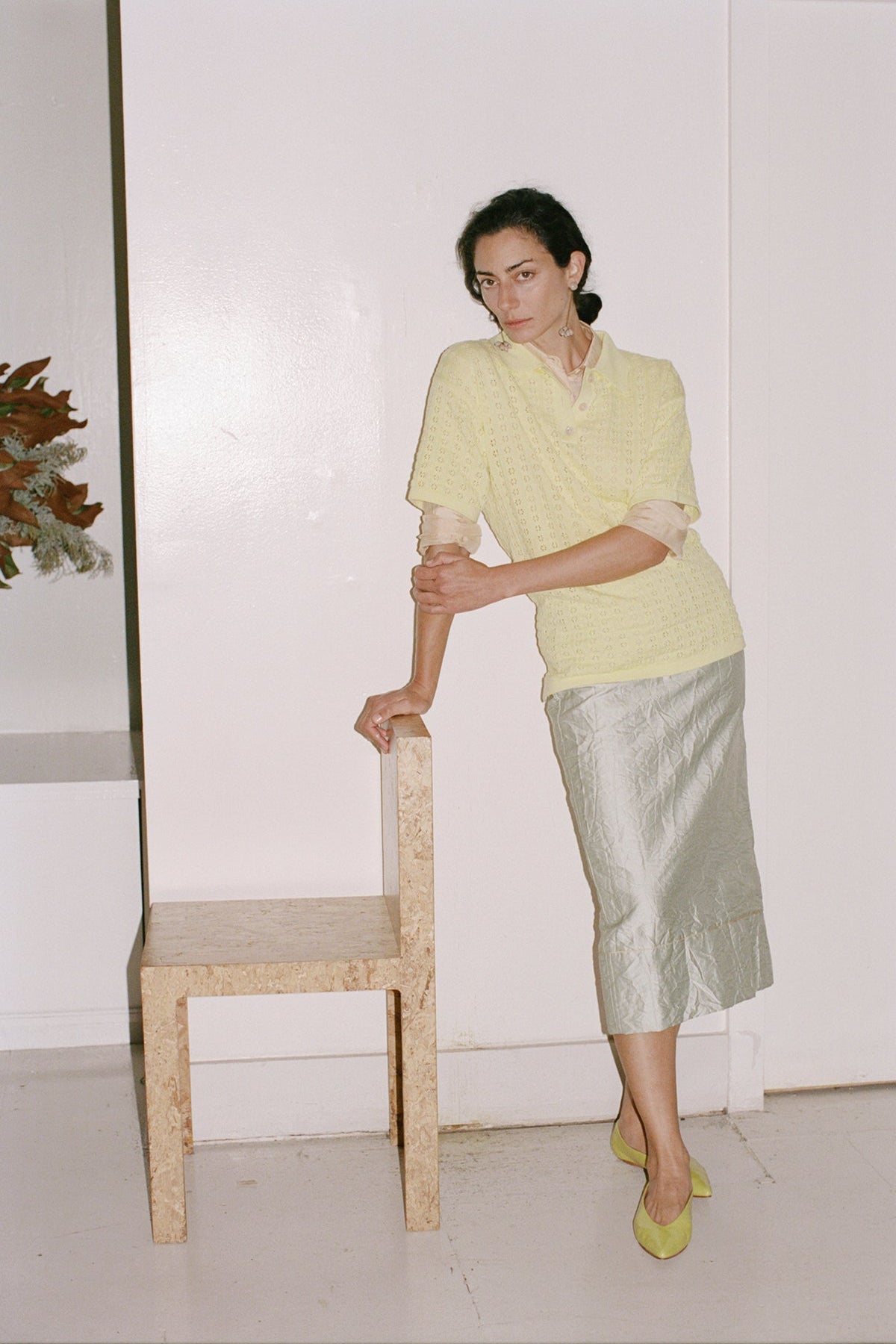 Woman in a yellow top and silver skirt standing next to a wooden chair against a white wall.