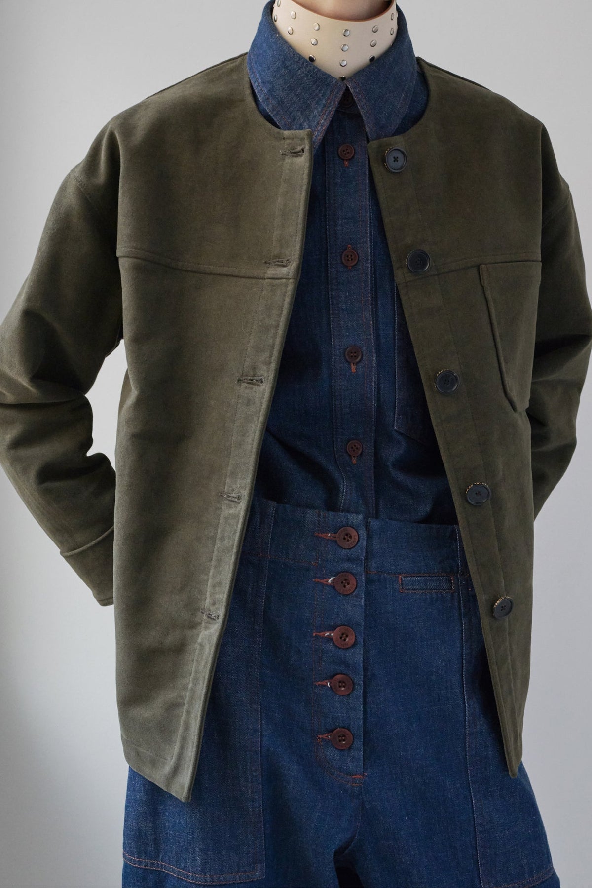 An olive green corduroy jacket styled over a blue denim shirt and matching denim overalls, shown on a mannequin against a neutral gray background.