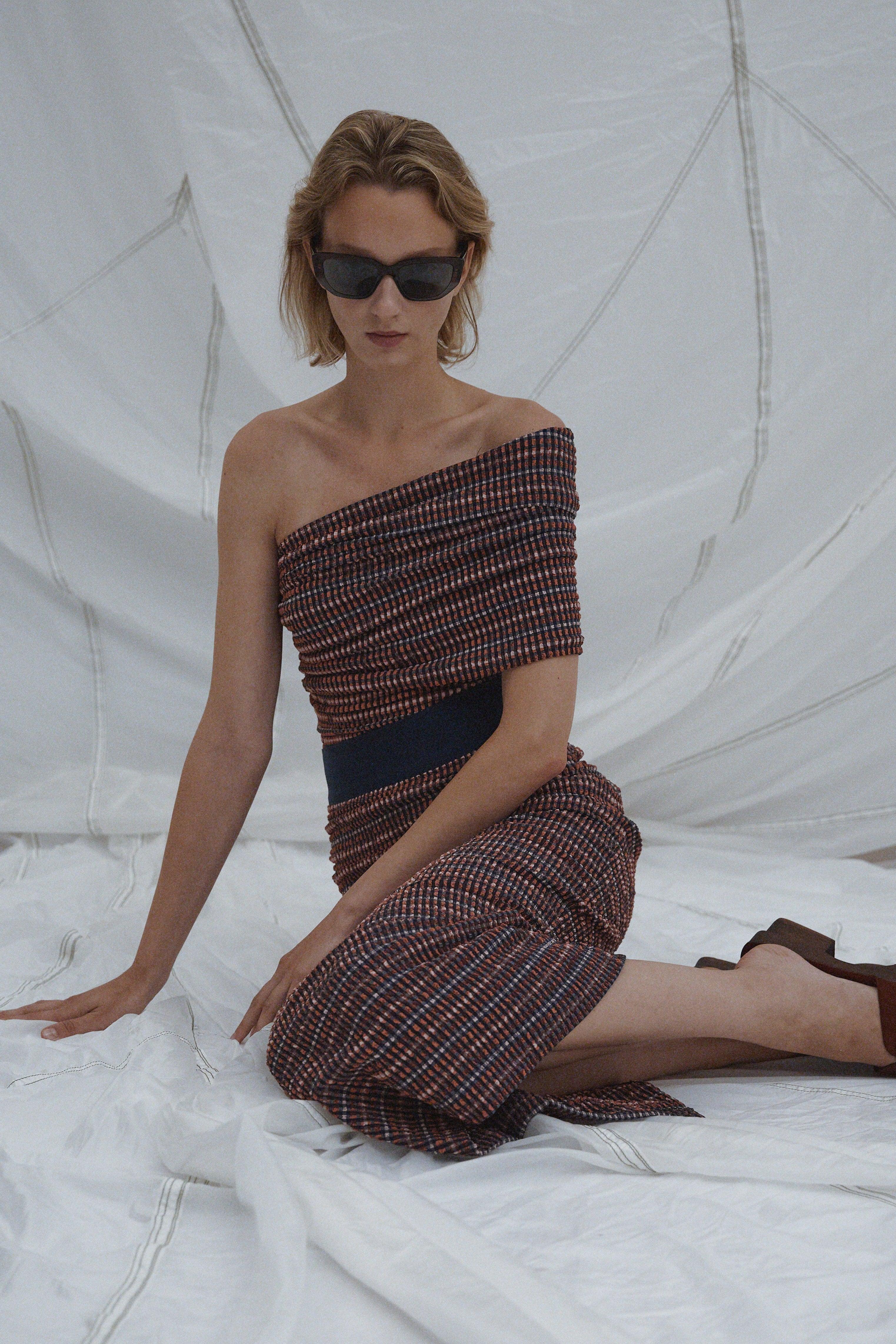 Woman in a Rachel Comey Spring 2026 patterned one shoulder top and matching skirt sitting on a white surface with a textured white background