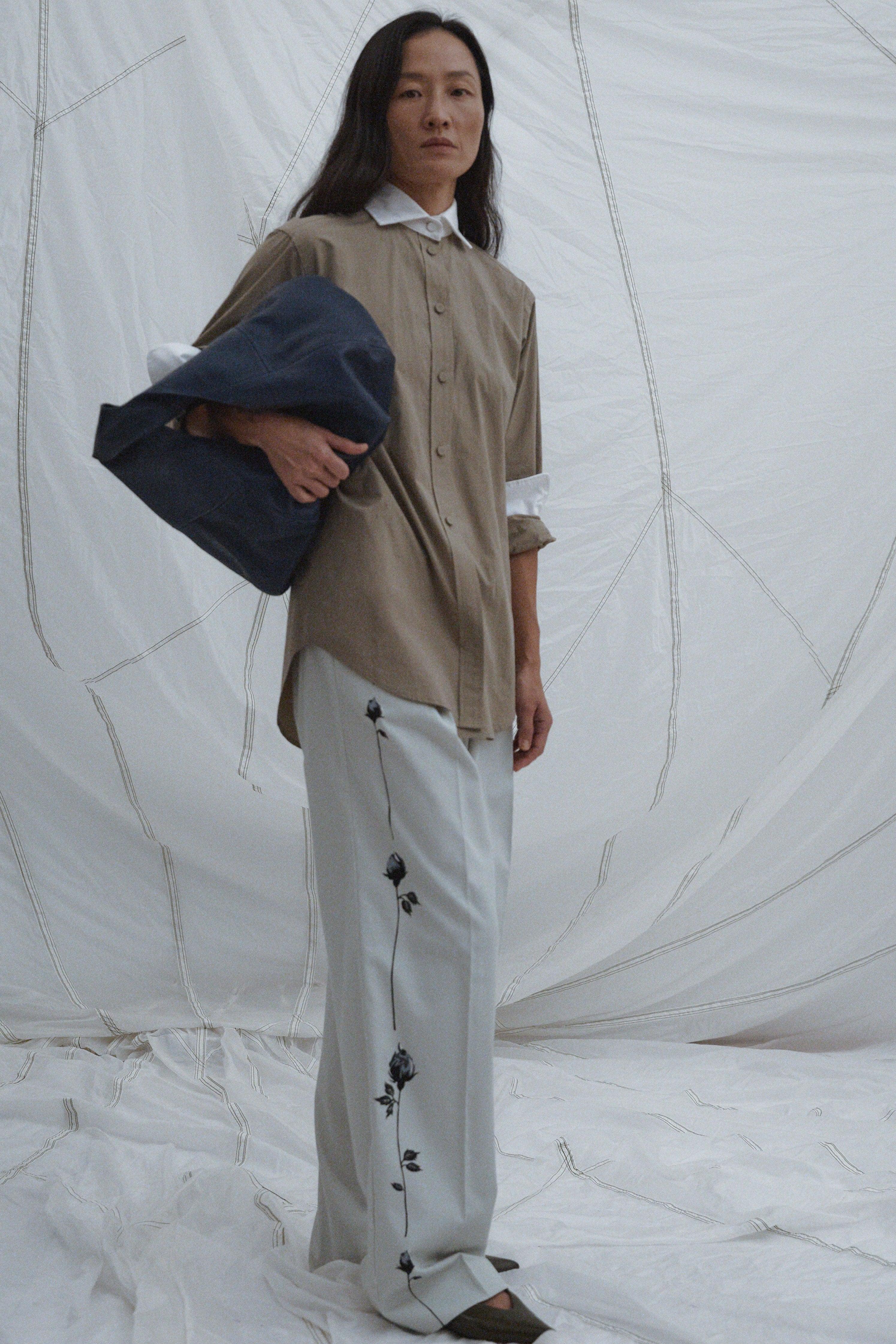 Person wearing a beige shirt and white pants with black floral patterns, holding a navy blue bag against a white background.