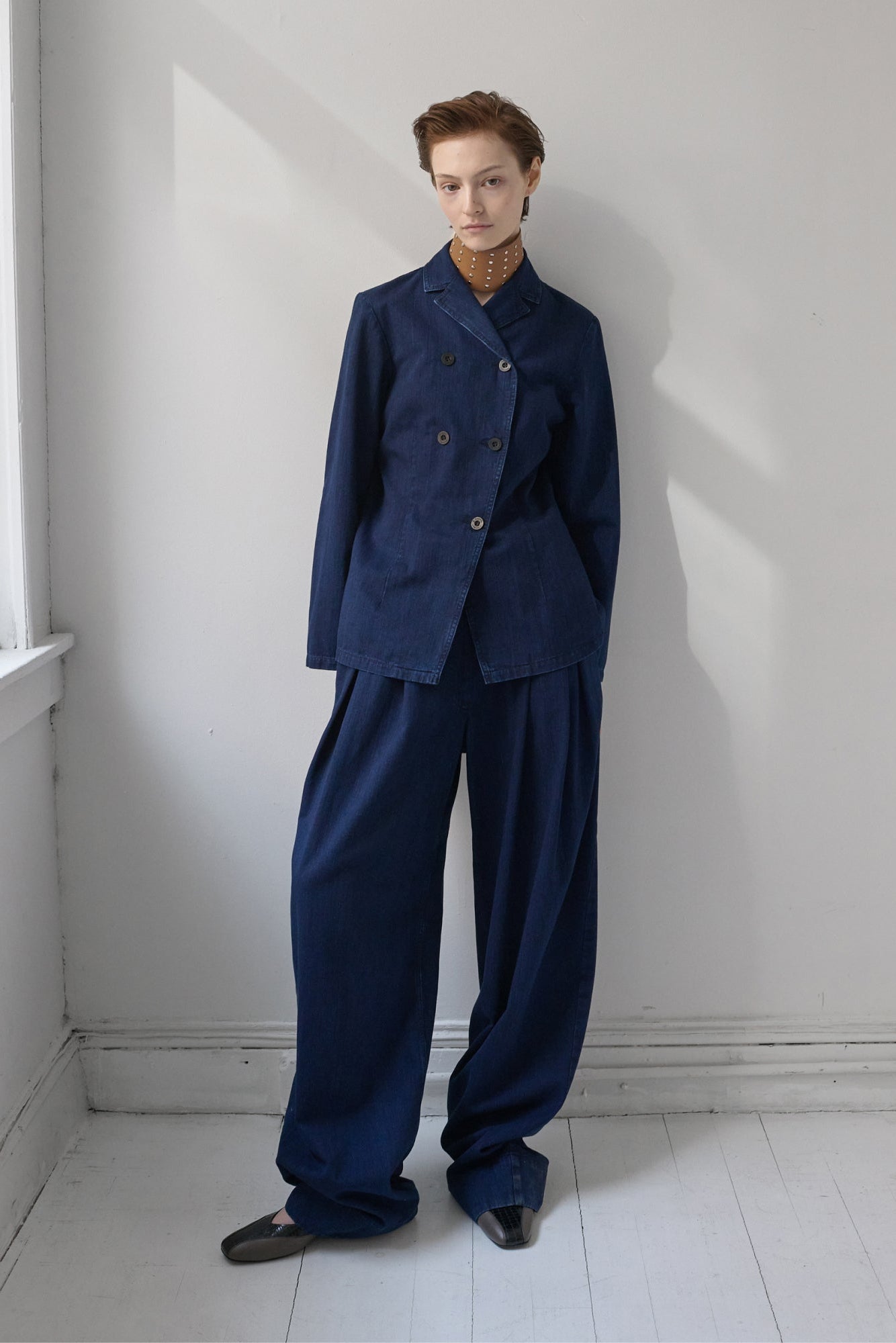 A model stands in a minimalist white room wearing a dark navy blue two-piece suit consisting of a double-breasted jacket and wide-leg trousers, layered over a brown necklace