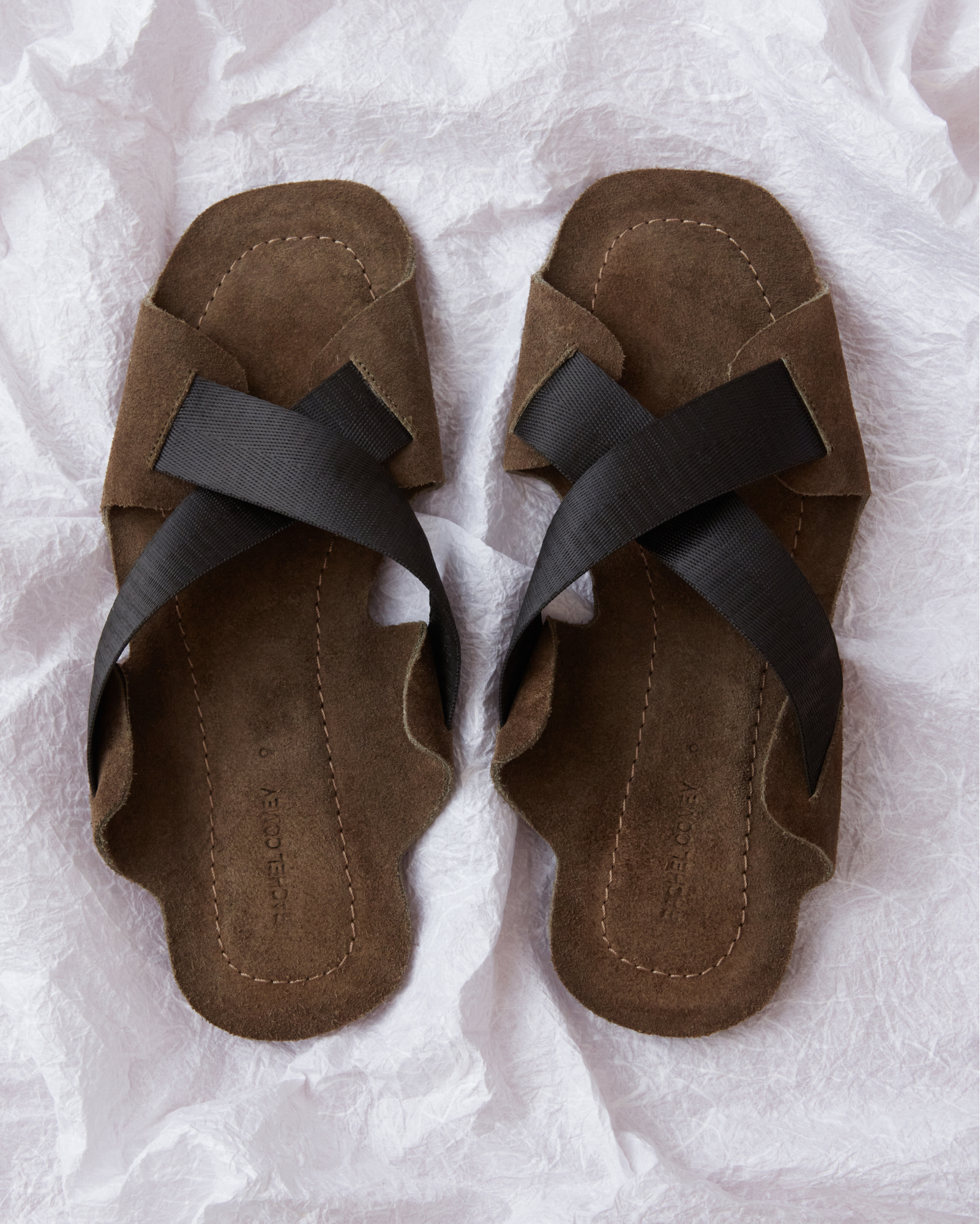 A pair of brown suede-like slide sandals with wide dark brown crisscross straps, laid flat on a crumpled white paper background.